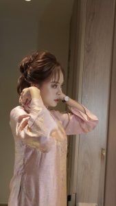 Phụ nữ thanh lịch chỉnh tóc trước gương với váy lụa hồng pastel, phong thái nhẹ nhàng và tinh tế